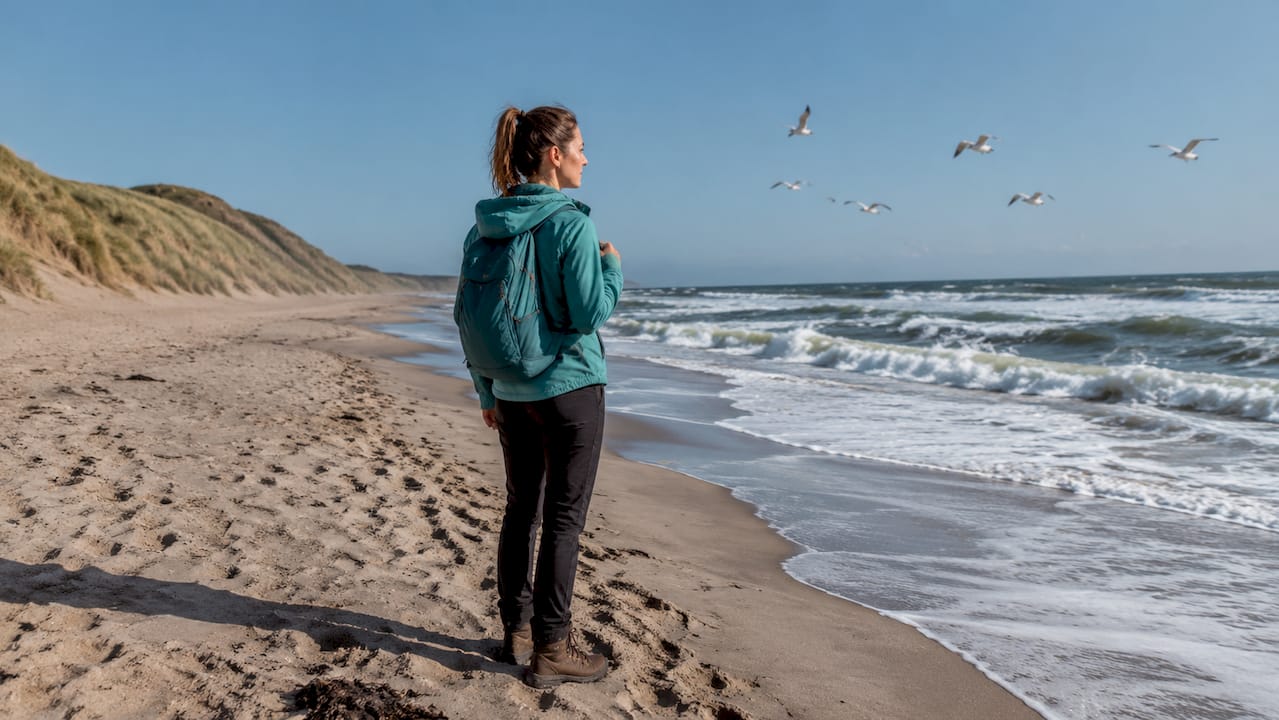 En kvinde går langs den blæsende strand ved Vestkysten, mens vinden rusker i hendes hår.
