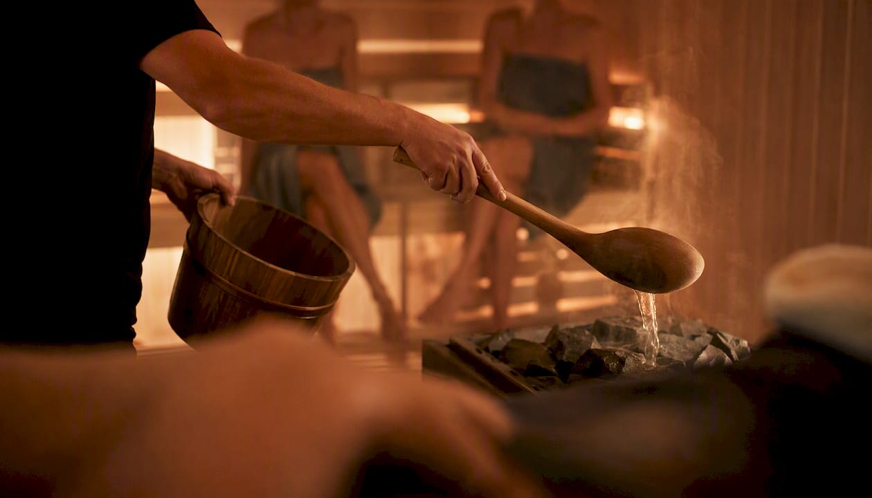 Eine Hand gießt Wasser über die heißen Steine der Sauna während einer Saunasitzung